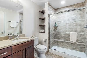 A modern bathroom with sliding glass door shower, built in towel shelves, toilet, and sink with cabinets