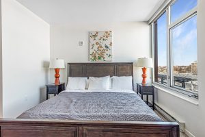 A bedroom with a large bed and night stands with lamps on either side on an upper level of an apartment building