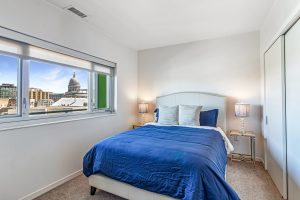 A bedroom with a large bed and night stands with lamps on either side on an upper level of an apartment building with a view of the Madison, WI capitol building out the window