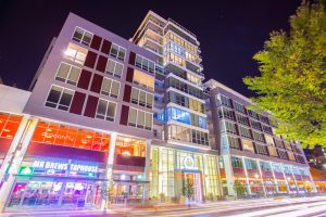 Exterior of a tall, glass building at night, with many bright neon lights at street level