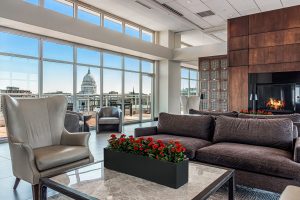 An apartment common room on the upper floor of a building with chairs, couches and a gas fireplace, and a view of the Madison, WI capitol building out the large windows